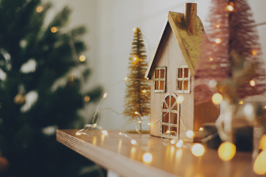 Miniature gingerbread house with lit windows surrounded by decorative trees and bokeh lights on wooden surface