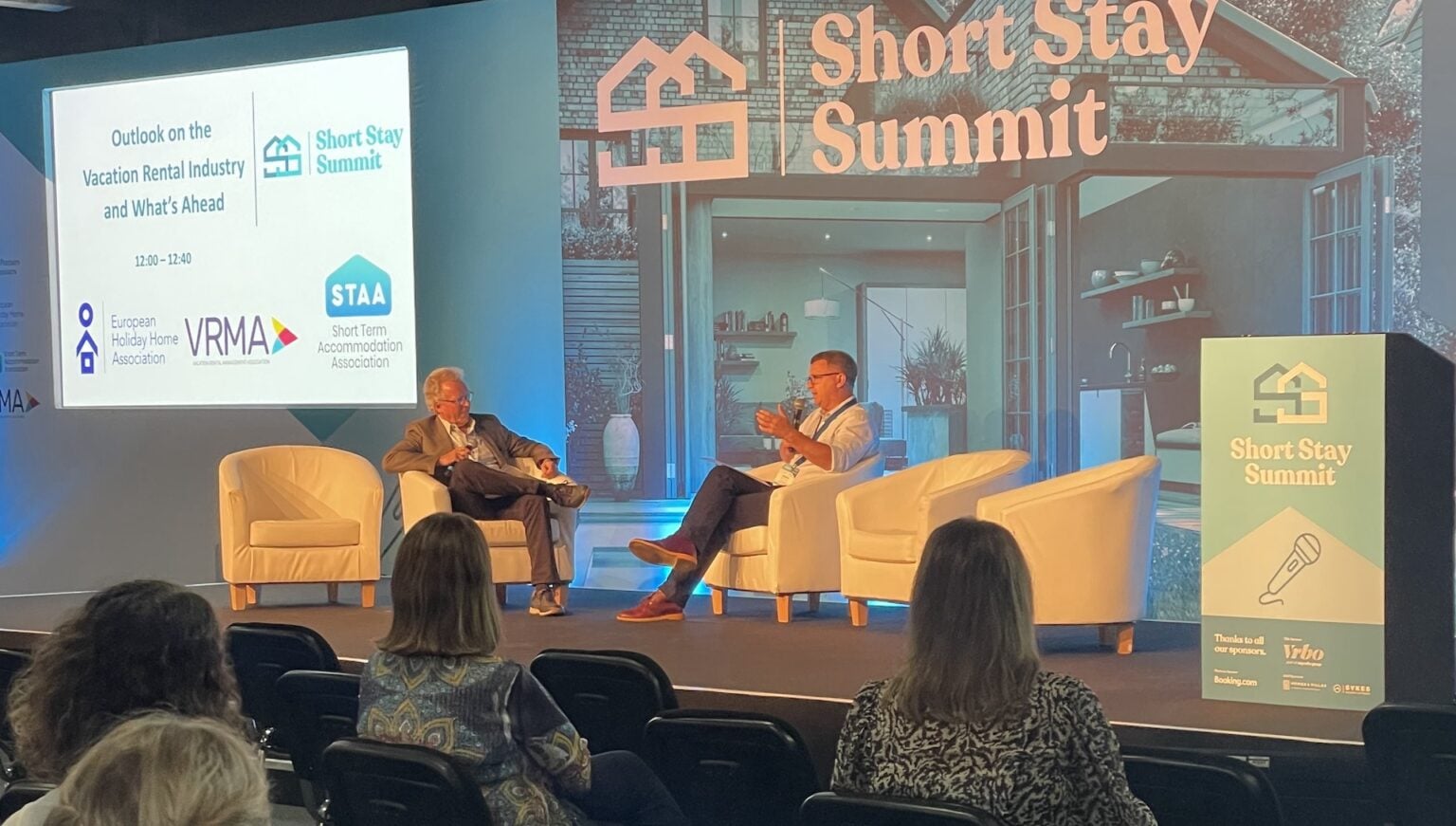 Two speakers in conversation on stage at Short Stay Summit, with audience members visible in foreground and presentation screen displaying event branding.