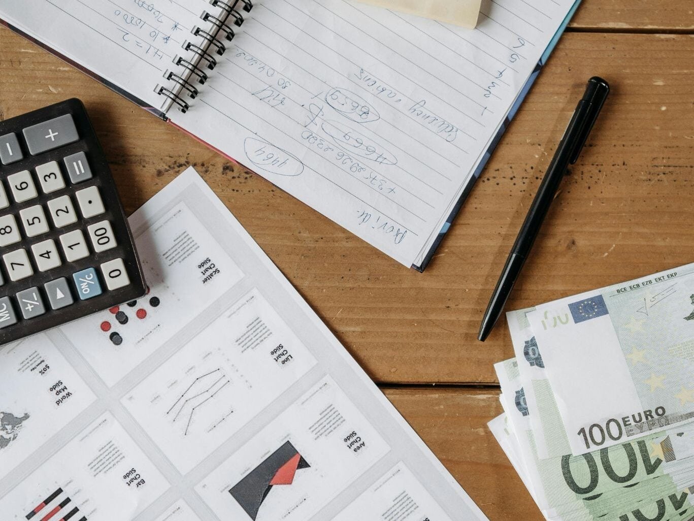 Workspace with financial documents, calculator, euro currency, sticky note on spiral notebook, and pen on wooden desk