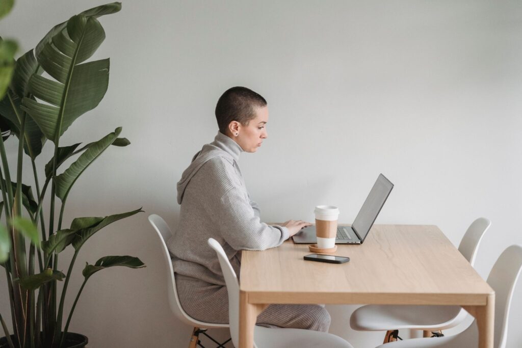 Professional working at laptop and coffee cup at wooden desk with potted plant in minimalist office setting