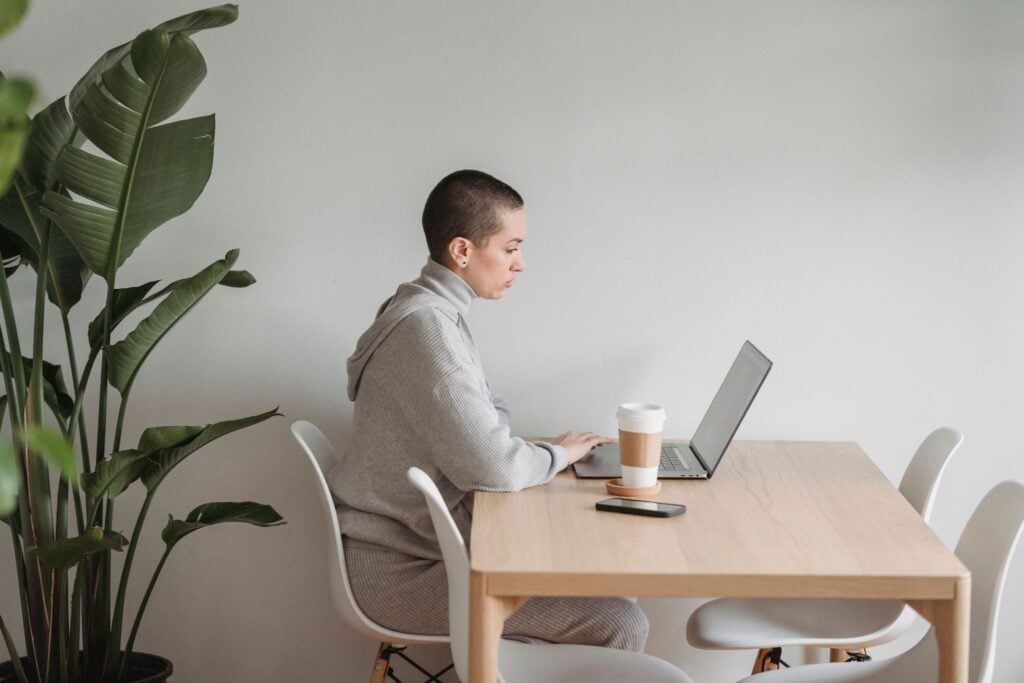 Person working on laptop at wooden desk with coffee cup and smartphone nearby, green plant in background
