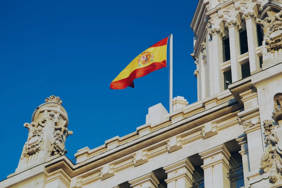 Bandera de España ondeando frente a edificio clásico con ornamentación arquitectónica bajo cielo azul
