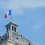 French flag flying above government building dome against blue sky