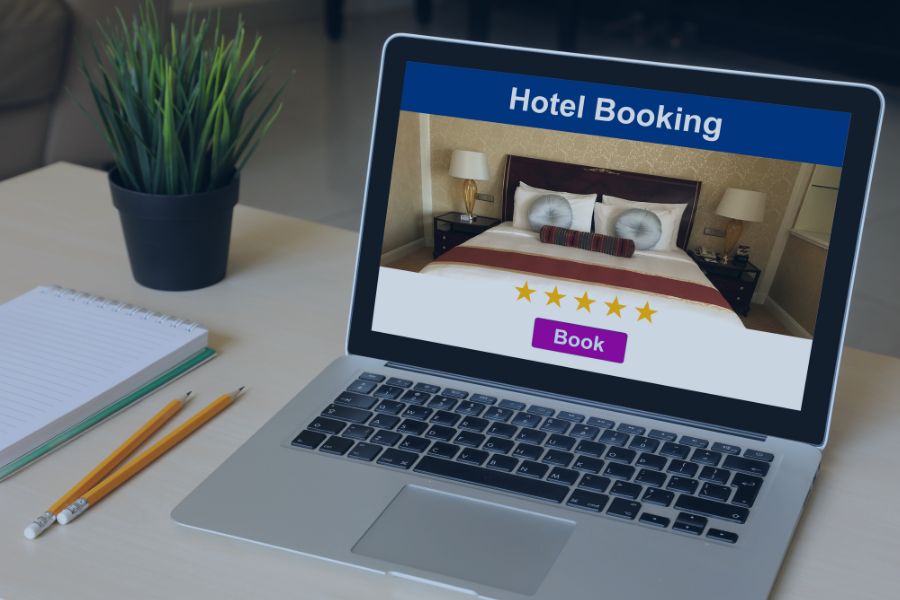 Laptop displaying hotel booking website with four-star rated bedroom and purple Book button, on desk with notepad and pencils
