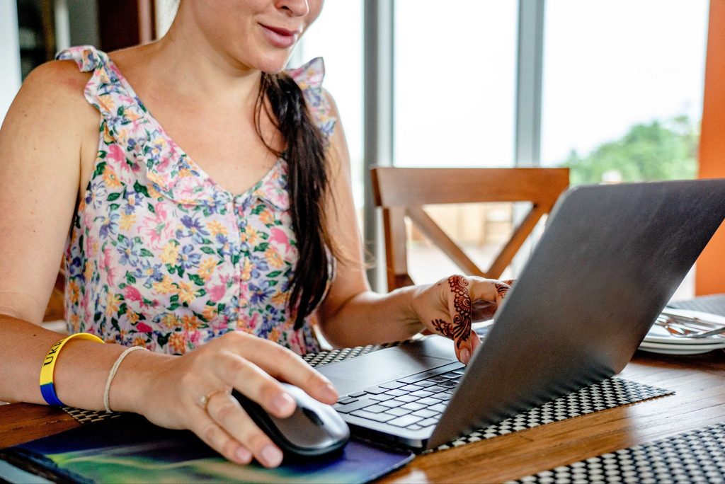 Mulher a trabalhar num laptop junto a uma janela, com rato sem fios e pulseira colorida visível