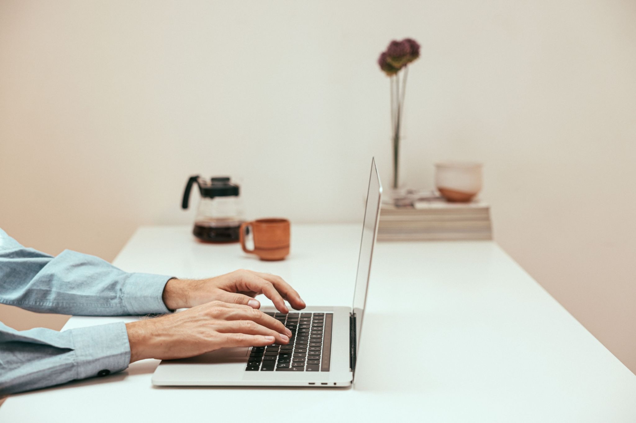 Person typing on laptop at white desk with coffee mug, camera, and dried flower in background